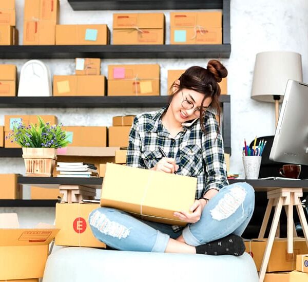 A woman sitting among boxes, talking on the phone, and writing on a package.